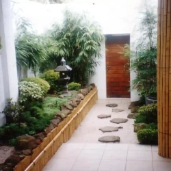 Entrance garden with bamboo wall cladding on low wall and on pillar. Japanese lantern and tsukubai water basin. Stones, rocks, gravel.
