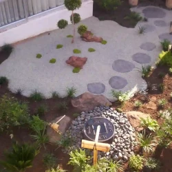 Japanese water basin (tsukubai) with bamboo water feature (kakehi), bamboo ladle and rest. Black modern Eastern lantern. Rocks. Gravel. Pebbles. Black slate stepping stones. Multi-head shaped tree. Wooden screen behind.