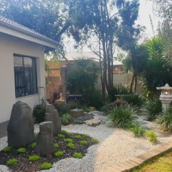 Three grey monolith rocks set into a mossy mound, surrounded by gravel, near the Japanese planted garden with a Japanese water basin, kakehi water feature, bench and Japanese lantern.