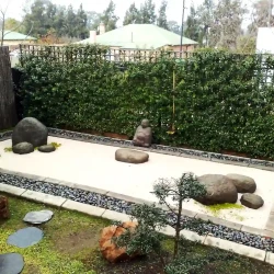 Path of black slate stepping stones leading from Japanese Zen raked garden with black pebble border into planted garden with shaped trees.