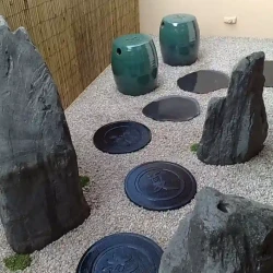 Tiny balcony garden. Path of decorative black round steppingstones, set into gravel leading to a small self-contained Zen water feature. Three monolit rocks, a small potted nandina, very few mossy ground covers and split bamboo cladding to add a finishing touch and texture. 2 tiny ceramic seats/tables