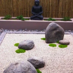 Dry raked Zen Japanese garden, rocks, mosses, raked gravel. Raised Buddha statue in planter with cascading junipers and wooden screen as background.